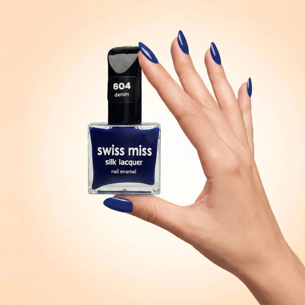 Swiss Miss Silk Lacquer Nail Enamel in shade Denim 604 held by hand showing clear glass square bottle with deep royal navy blue polish and matching manicured nails.