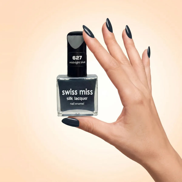 Swiss Miss Silk Lacquer Nail Enamel in shade Midnight Blue 627 held by hand showing clear glass square bottle with deep midnight blue polish and matching manicured nails.