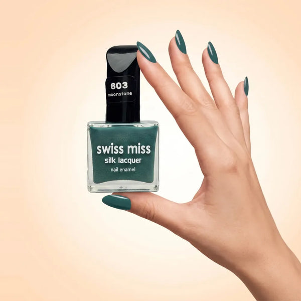 Swiss Miss Silk Lacquer Nail Enamel in shade Moonstone 603 held by hand showing clear glass square bottle with deep teal green polish and matching manicured nails.