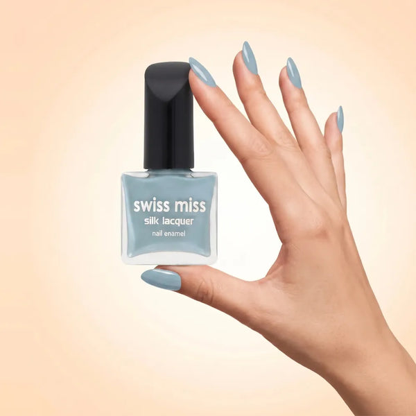 Swiss Miss Silk Lacquer Nail Enamel in shade Raindrop 631 held by hand showing clear glass square bottle with dusty muted sky blue polish and matching manicured nails.