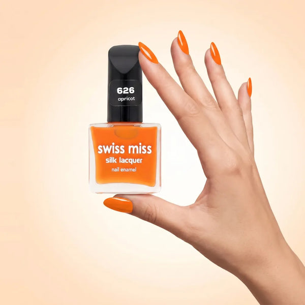 Swiss Miss Silk Lacquer Nail Enamel in shade Apricot 626 held by hand showing clear glass square bottle with bright apricot orange polish and matching manicured nails.