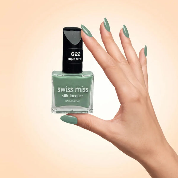 Swiss Miss Silk Lacquer Nail Enamel in shade Aqua Forest 622 held by hand showing clear glass square bottle with soft sage aqua green polish and matching manicured nails.