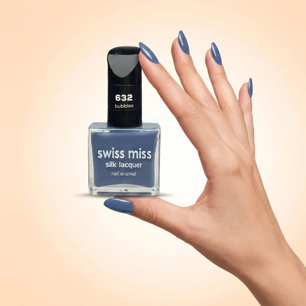 Swiss Miss Silk Lacquer Nail Enamel in shade Bubbles 632 held by hand showing clear glass square bottle with dusty slate denim blue polish and matching manicured nails.
