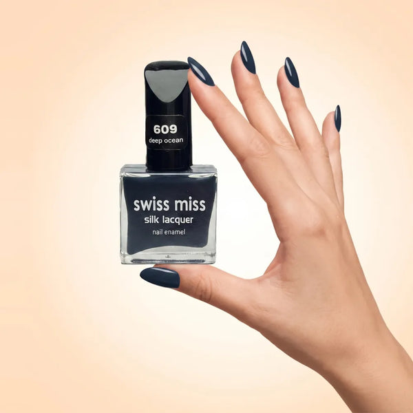 Swiss Miss Silk Lacquer Nail Enamel in shade Deep Ocean 609 held by hand showing clear glass square bottle with dark ocean navy blue polish and matching manicured nails.