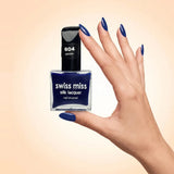 Swiss Miss Silk Lacquer Nail Enamel in shade Denim 604 held by hand showing clear glass square bottle with deep royal navy blue polish and matching manicured nails.