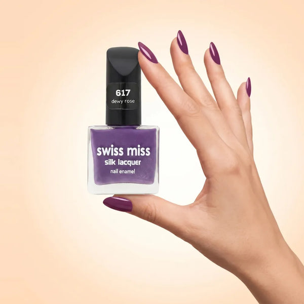Swiss Miss Silk Lacquer Nail Enamel in shade Dewy Rose 617 held by hand showing clear glass square bottle with deep plum mauve purple polish and matching manicured nails.