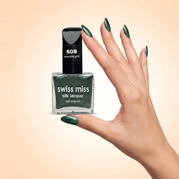 Swiss Miss Silk Lacquer Nail Enamel in shade Emerald Gold 608 held by hand showing clear glass square bottle with metallic shimmer forest emerald green polish and matching manicured nails.