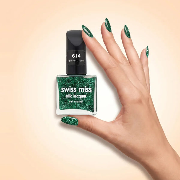 Swiss Miss Silk Lacquer Nail Enamel in shade Glitter Green 614 held by hand showing clear glass square bottle with sparkling emerald green glitter polish and matching manicured nails.
