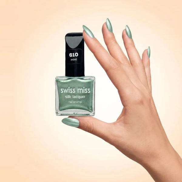 Swiss Miss Silk Lacquer Nail Enamel in shade Mint 610 held by hand showing clear glass square bottle with metallic shimmer light mint green polish and matching manicured nails.
