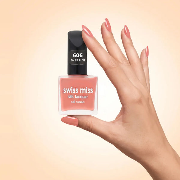 Swiss Miss Silk Lacquer Nail Enamel in shade Nude Pink 606 held by hand showing clear glass square bottle with soft peachy nude pink polish and matching manicured nails.