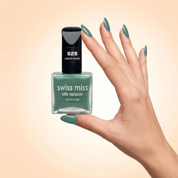 Swiss Miss Silk Lacquer Nail Enamel in shade Pastel Dream 628 held by hand showing clear glass square bottle with muted pastel sage mint green polish and matching manicured nails.