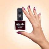 Swiss Miss Silk Lacquer Nail Enamel in shade Plump 602 held by hand showing clear glass square bottle with deep plum berry polish and matching manicured nails.