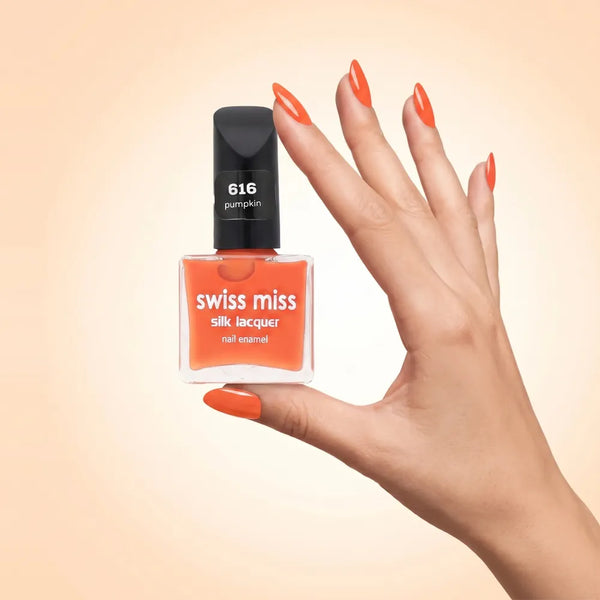 Swiss Miss Silk Lacquer Nail Enamel in shade Pumpkin 616 held by hand showing clear glass square bottle with bright vibrant pumpkin orange polish and matching manicured nails.
