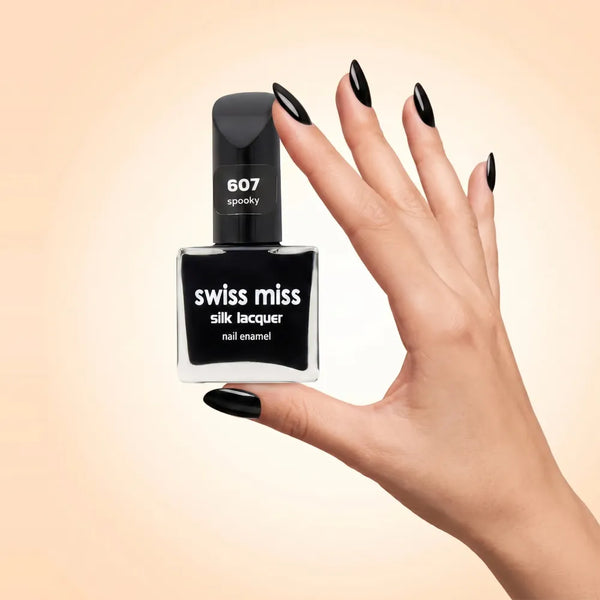 Swiss Miss Silk Lacquer Nail Enamel in shade Spooky 607 held by hand showing clear glass square bottle with deep jet black polish and matching manicured nails.