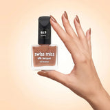 Swiss Miss Silk Lacquer Nail Enamel in shade Tawny 613 held by hand showing clear glass square bottle with warm light tawny brown nude polish and matching manicured nails.