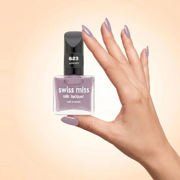 Swiss Miss Silk Lacquer Nail Enamel in shade Unicorn 623 held by hand showing clear glass square bottle with soft dusty lilac purple polish and matching manicured nails.