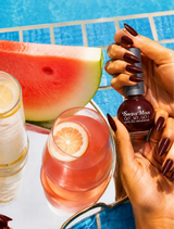 Woman holding Swiss Miss dark maroon NP Teracotta 208 quick dry nail polish by a pool with matching burgundy manicure.