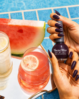 Woman holding Swiss Miss Get Set Go quick dry nail polish bottle in Deep Maroon 226 with a dark plum maroon manicure by a pool.