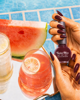 Woman holding Swiss Miss Get Set Go quick dry nail polish bottle in Ribbon Red 224 with a dark cherry red manicure by a pool.