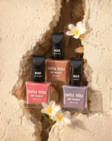 Three bottles of Swiss Miss Silk Lacquer nail enamel framed in a textured stone wall crack adorned with white plumeria flowers.